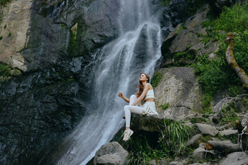 Woman meditating near waterfall dressed in white activewear sitting peacefully on rocks surrounded...