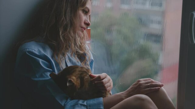 Unhappy beauty woman crying looking at window rain drops falling dark rainy weather, mood depression sadness loneliness emotion at home. Adult upset Girl sad tear-stained face eyes. hand holds pet cat