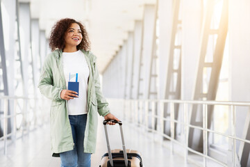 Happy African American woman with travel documents and suitcase walking forward in airport...