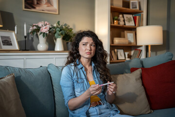 Worried woman looking at pregnancy test sitting on sofa at home