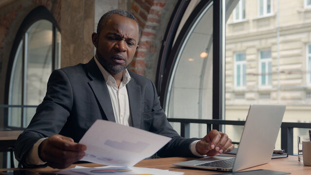 African American entrepreneur typing laptop computer check data in papers dissatisfied problem mistake report bad result sad stress business man office businessman male guy research analyse documents