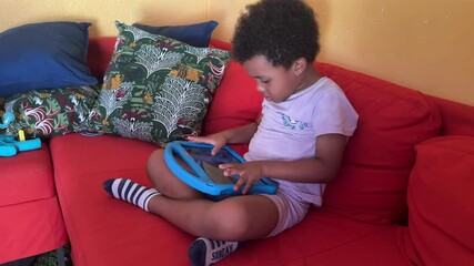 5-year-old african black child playing with his tblet sitting on a red coach at home. He is wearing curly hair and summer chothing. - Powered by Adobe