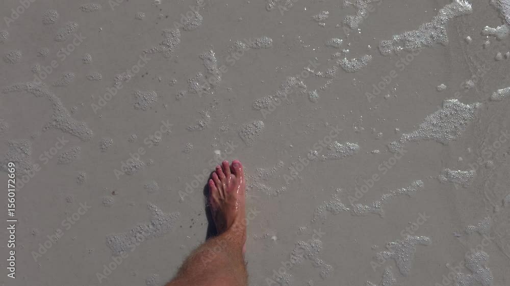 Poster a barefoot person slowly steps across smooth wet sand as gentle ocean foam washes the shore. the slo