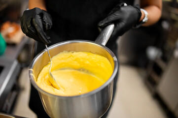 Chef wearing black gloves stirring creamy yellow sauce in stainless steel pot, illustrating...