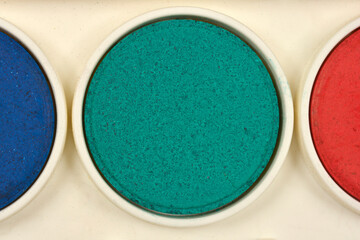 A close-up shows three oval pans of dry watercolor paint in blue, green, and red within a white...
