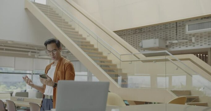 Senior Asian businessman wearing headset celebrates success during online meeting in modern co-working space. Remote work and business communication concept.