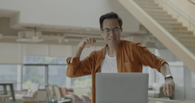Senior Asian businessman wearing headset celebrates success during online meeting in modern co-working space. Remote work and business communication concept.