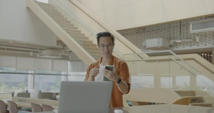 Senior Asian businessman wearing headset celebrates success during online meeting in modern co-working space. Remote work and business communication concept.