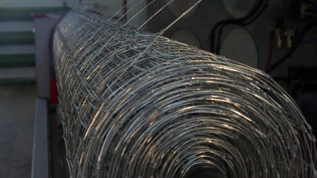 A close-up view of a large, tightly wound roll of galvanized wire mesh, likely a fixed-knot fence, sitting on an industrial machine.