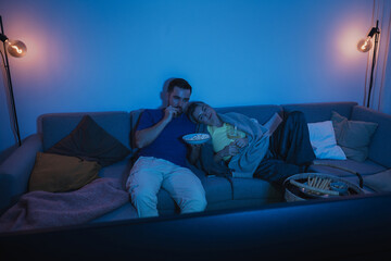Couple watching TV at night  while sharing a bowl of popcorn and wine on sofa
