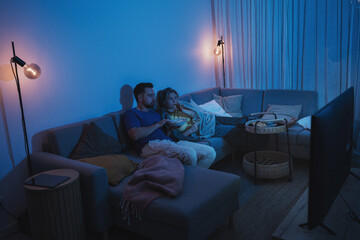 Couple watching TV at night  while sharing a bowl of popcorn and wine on sofa