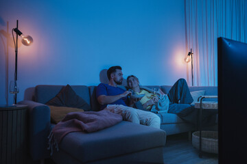 Couple watching TV at night  while sharing a bowl of popcorn and wine on sofa