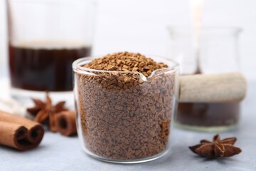 Granulated instant coffee in measuring cup and spices on grey table against light background, closeup