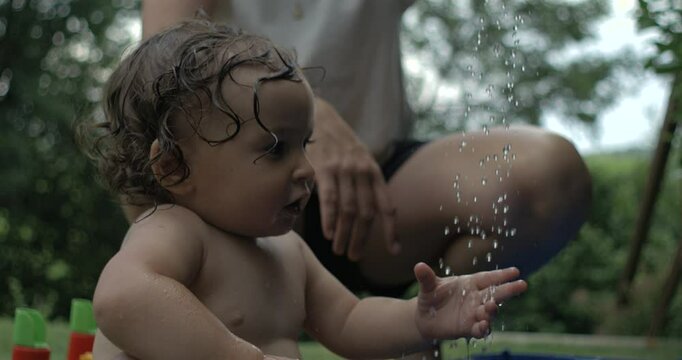 Curious baby with wet hair reaching out toward falling water droplets in slow motion, adult figure in background, surrounded by green outdoor scenery