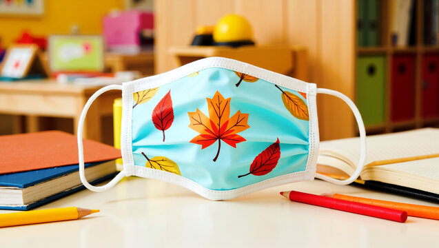 Child's face mask with autumn leaf pattern on a table with school supplies, representing back to school during pandemic, child safety, and seasonal themes in a classroom setting.