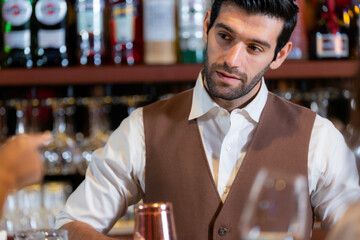 Close-up portrait of a handsome, attentive young bartender with a beard, wearing a vest and listening to an order at a classic bar. Professional service in a restaurant.