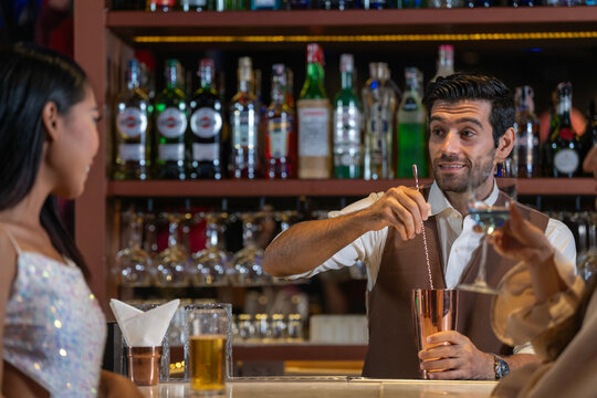 Expatriate bartender skillfully preparing cocktails for two beautiful female customers at a stylish bar counter in Bangkok, Thailand. Warm atmosphere and friendly service in a vibrant city.
