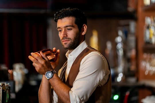 A handsome and focused professional bartender concentrating as he prepares a craft cocktail with a copper shaker in a stylish, dark bar at night.
