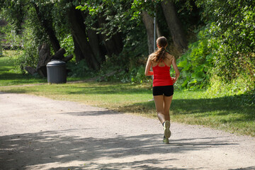 A sporty young woman in a red top and black shorts jogging in a city park. Active lifestyle in modern city