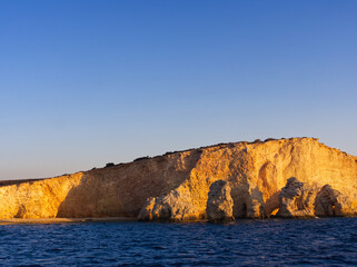 Sunlit golden limestone cliffs rising from calm blue waters, with clear sky above, at golden hour. Koufonisia Islands, Cyclades, Greece