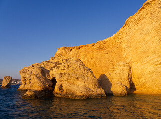 Dramatic golden limestone cliffs and rock formations rising from calm blue waters under clear sky....