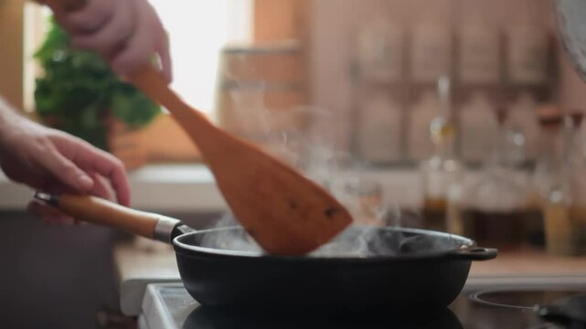 A man adds chopped celery to a hot, steaming pan on the stove. Spices and fresh herbs are blurred in the background. Concept of home cooking