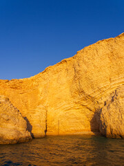 Stunning golden limestone cliffs towering over tranquil blue waters, illuminated by warm sunset light. Koufonisia Islands, Cyclades, Greece