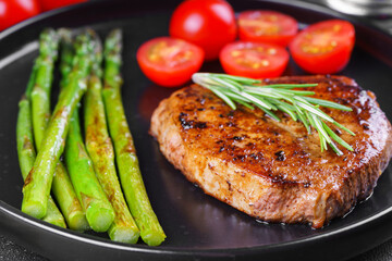 Close up, macro of tasty grilled beef steak served with green asparagus and cherry tomatoes on black plate, with cutlery. Perfect for food menu, diet, or culinary design concepts.