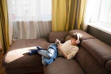  preteen boy wearing headphones listens to music and watches a video sitting on the couch at home....