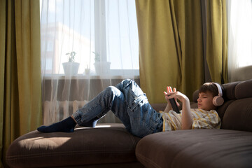  preteen boy wearing headphones listens to music and watches a video sitting on the couch at home....