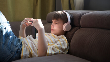  preteen boy wearing headphones listens to music and watches a video sitting on the couch at home. favorite music. Generation Z.  © ulza