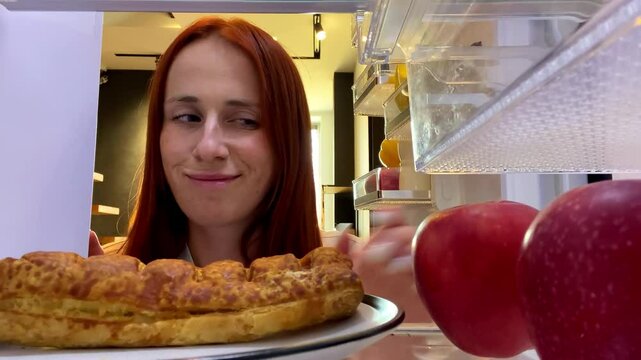 View from inside refrigerator hungry woman at kitchen taking apple over sweet bun dessert,healthy nutrition,diet,weight control,obesity overeating food addiction