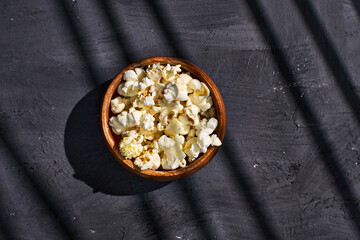 A wooden bowl filled with fluffy, freshly popped popcorn rests on a dark textured surface