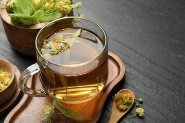 Tasty linden tea in cup, spoon, leaves and flowers on dark textured table, closeup