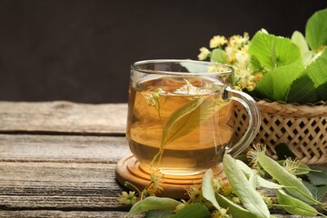 Tasty linden tea in cup, leaves and flowers on wooden table against dark background, closeup. Space...