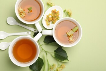 Tasty linden tea in cups, spoons, leaves and flowers on light green background, flat lay. Space for text