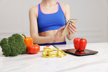 Woman using smartphone at white marble table with kitchen scale, vegetables and measuring tape...