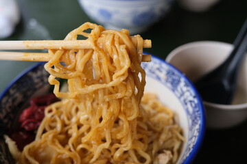 Close-up of chopsticks picking up yellow, seasoned noodles from a decorative bowl, part of a traditional Asian noodle dish.