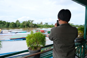 A person takes a photo of colorful wooden boats on a tropical river from a balcony, capturing the...