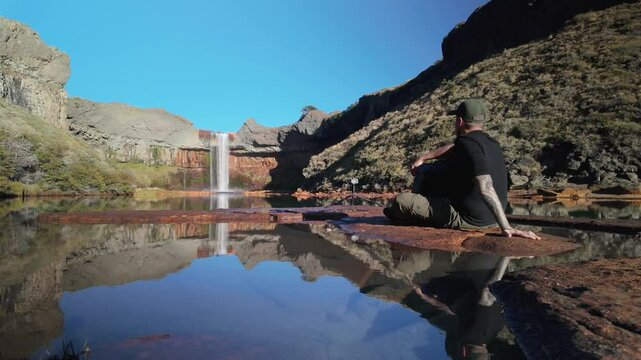Man sitting at the base of the Salto del Agrio waterfall and reflected pond and rocky cliffs. Neuquen, Patagonia Argentina