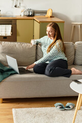 Cozy moments of a young woman enjoying her home space while using a laptop and sipping tea