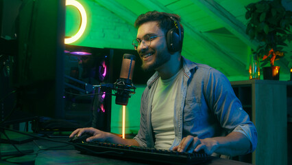 Emotional smiling talking in microphone video game addict Caucasian guy male man streamer gamer wearing headphones playing virtual battle internet entertainment web online carefree computer at night