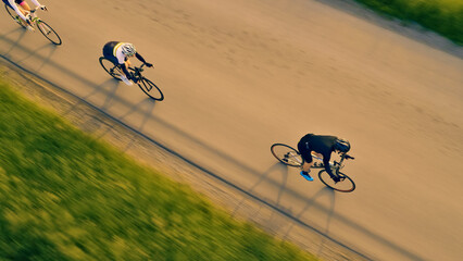 A group of cyclists pedal down a road, captured in an aerial view. The motion blur emphasizes their...