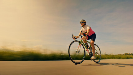 A woman cyclist rides her bike on an open road, wearing a helmet and sunglasses. The background features a blurred landscape, emphasizing speed and focus. Aerial view.