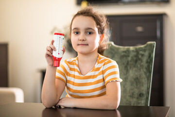 Cute little girl monitoring peak expiratory with peak flow meter at home. Hand medical device used...