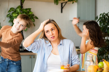 Young woman mom sitting on a kitchen table looking stressed suffers from headache because of noise...