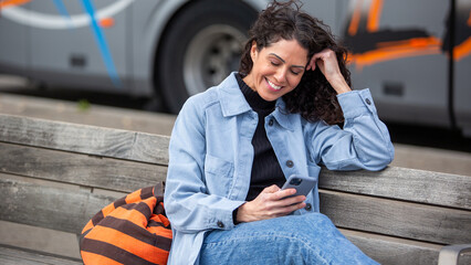 young woman smiling while using her smartphone on a bench, with a travel bag beside her and a bus in the background.