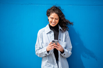 Young woman smiling while using her phone, standing against blue wall with the wind blowing through...