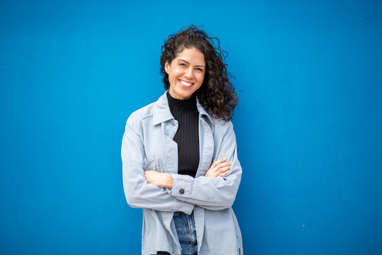 Confident woman with curly hair standing against blue wall, smiling with arms crossed in a casual and relaxed pose.