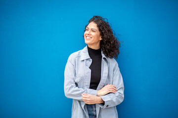 Smiling woman with curly hair standing confidently against a bright blue wall, dressed in casual...